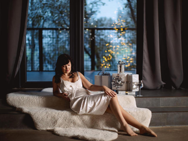 NathalyStars with tits in a white satin dress, lounging on a fluffy rug, legs crossed, near a window with Christmas lights outside, and wrapped presents on a windowsill.