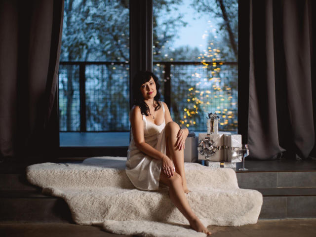 NathalyStars sits on a white furry rug, wearing a white dress, showing her tits and ass, with dark hair, hands on her knee, and a gift box beside her.