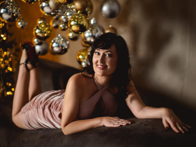 NathalyStars laying on a couch, wearing a pink, criss-cross dress, black heels, smiling, with her ass up, and a cluster of shiny ornaments in the background.