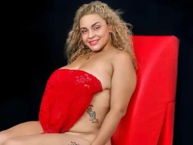 MarieRouxPassion with curly blonde hair, red lace strapless top covering her boobs, sitting on a red chair, smiling, tattoo on her right side, black background.