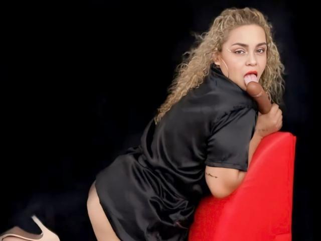MarieRouxPassion, with curly blonde hair, black satin dress, and high heels, leans over a red chair, licking a chocolate dildo, showing her bare ass.