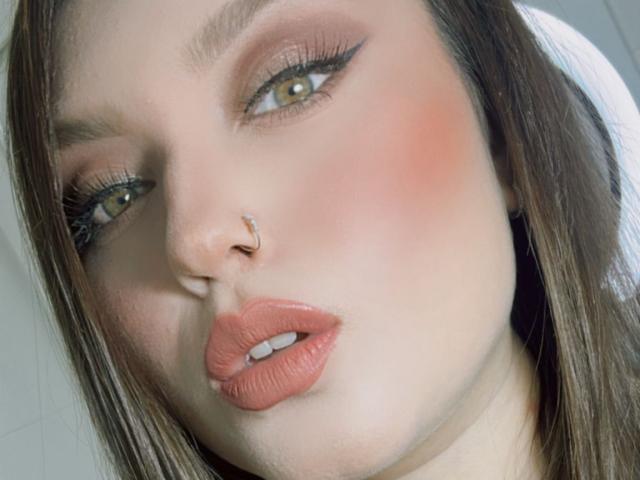 LillithDeluxe's face with light brown eyes, pink blush, rose-colored lips, nose ring, and long brown hair, looking slightly up with parted lips showing a small bit of her teeth.