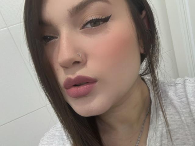 LillithDeluxe has a cute face with pink lips, brown eyes, and thick black eyeliner. She has a nose piercing and long brown hair. She's wearing a gray shirt.