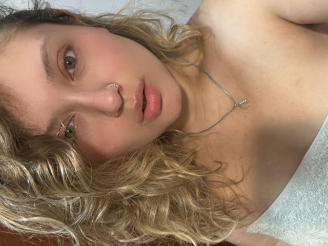 LillithDeluxe has curly blonde hair, a nose ring, and a necklace, with her pink lips slightly parted, showing her bare shoulders and a soft, pale skin tone.