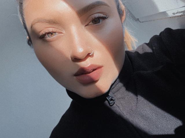 LillithDeluxe with light skin, brown eyes, and a nose ring, wearing a black zip-up, sunlight highlighting her face, slightly parted lips, and long eyelashes in a selfie.