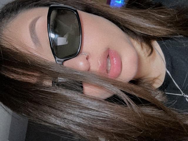 LillithDeluxe has long brown hair covering her eyes, wears black glasses, and has shiny pink lips with a small nose piercing. She's wearing a black top and a necklace.