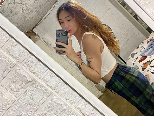 QueenAmaraTS, avec de longs cheveux bruns, un débardeur blanc et un pantalon à carreaux verts, prend un selfie devant un miroir, avec des tatouages visibles, tout en tenant un téléphone avec une coque bleue..