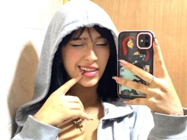 PriscilaLyn with black hair, grey hoodie, and finger in mouth, smiling, holding phone with a colorful case, wooden background. Boobs not visible, focus on face and phone.