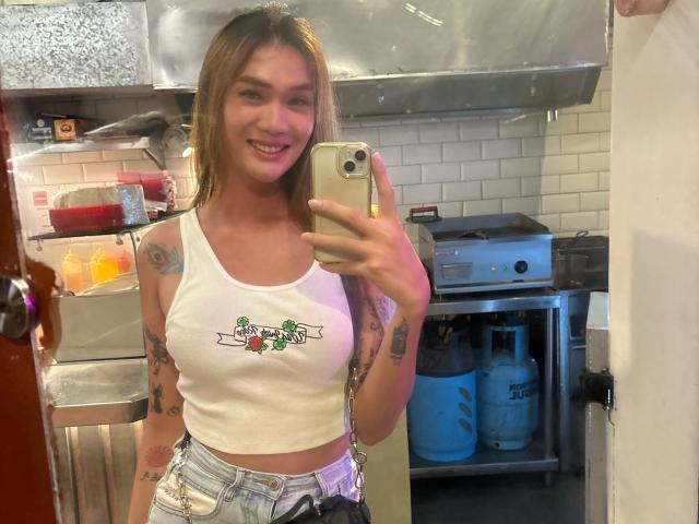 QueenAmaraTS with tattoos, smiling, wearing a white crop top showing small tits and denim shorts, taking a mirror selfie in a kitchen, holding an iPhone, with blonde hair.