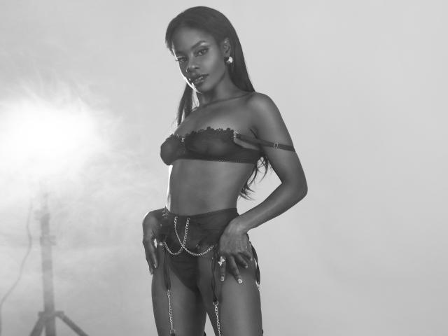 MiaHopson with dark skin, long black hair, wearing a black lace bra and matching high-waisted panties with chains, standing confidently, hands on hips, looking at the camera.