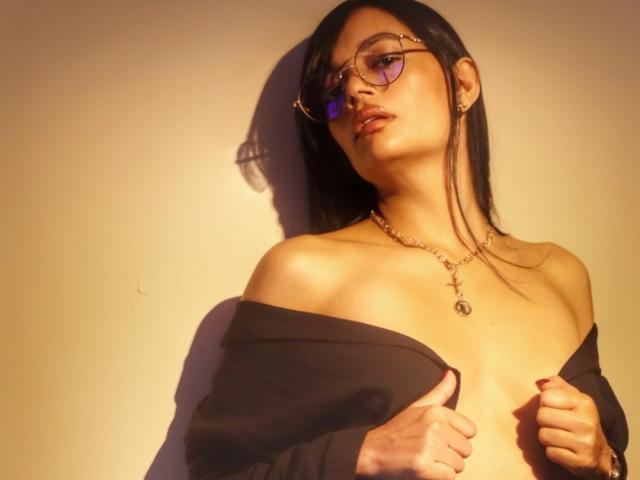 SthefanyGomez with glasses, black off-shoulder shirt, necklace, and a slight tan, holding her shirt to expose her tits, dark hair, and a sultry expression.
