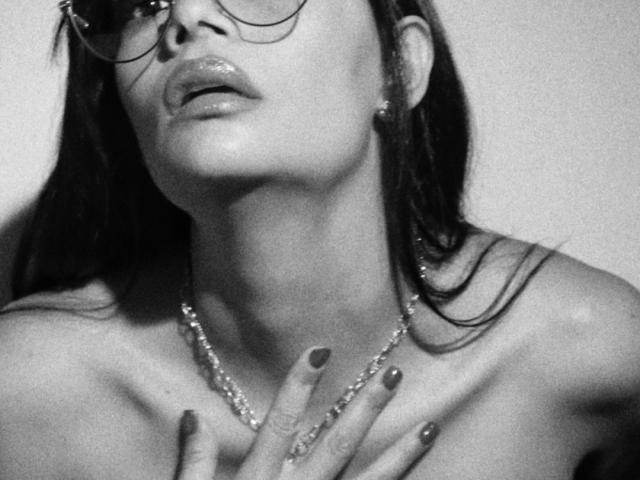 SthefanyGomez with glasses, glossy lips, and a necklace, her hand covering her tits, black nail polish, and a pearl earring, in a grayscale, slightly blurred photo.