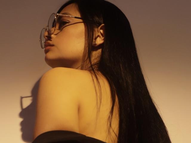 SthefanyGomez with long black hair, glasses, and a bare shoulder, looking to the side in warm, golden light, shadow on the left. Her ass is partially visible.