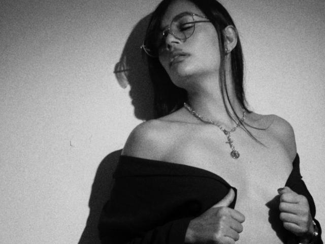 Black and white photo of SthefanyGomez with glasses, dark hair, necklace, off-shoulder shirt, showing bare chest, holding shirt, shadow on wall, eyes closed, looking sexy.