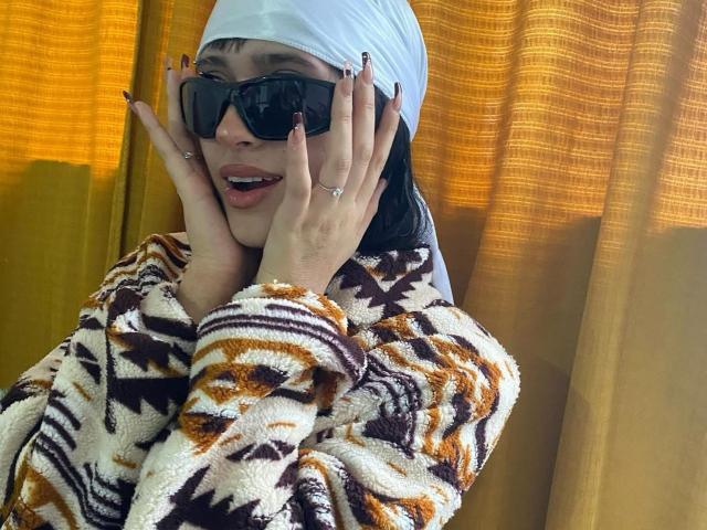 PriscilaLyn with black nails, white headscarf, big black sunglasses, smiling, hands on cheeks, wearing a brown and white patterned fuzzy sweater, golden-yellow curtain in background.
