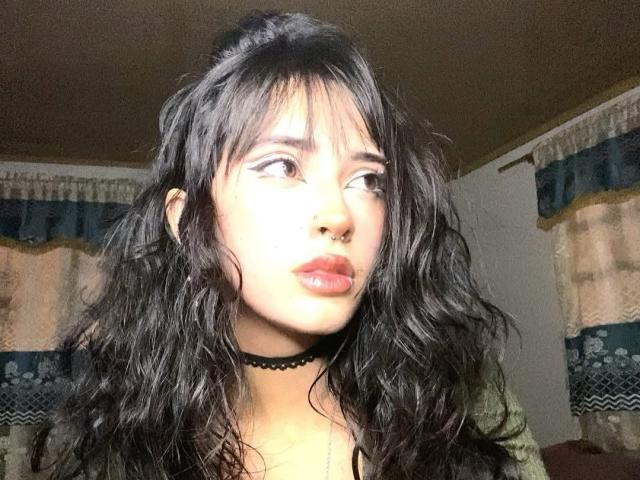 PriscilaLyn has wavy black hair, a nose ring, and a black choker. She's looking to the side with glossy red lips, wearing a green top.
