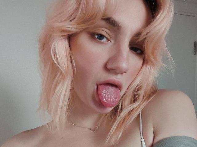 Arina, con cabello rosa, lengua afuera, mirando hacia arriba, con la boca ligeramente abierta, mostrando la superficie húmeda de su lengua, vistiendo un collar delgado y una blusa gris con un hombro descubierto..