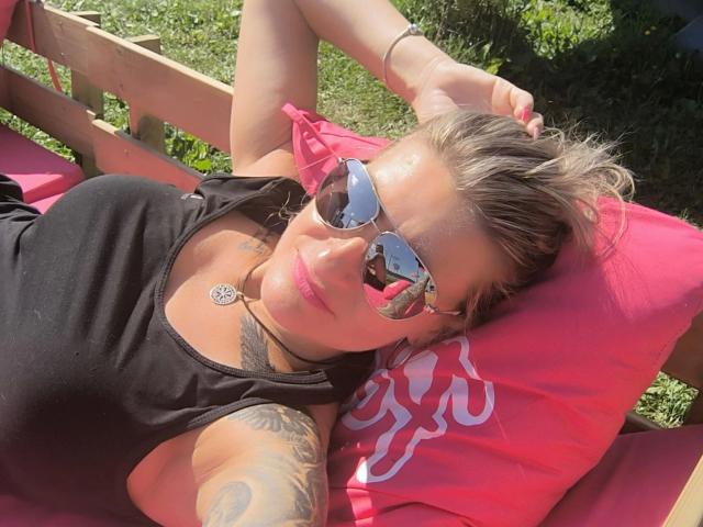 KatyStar-hot lounging on a pink pillow, wearing black top, reflective sunglasses, sunlit tattoos, arm raised, brown hair, pink lips, chest visible, grassy background, sunny day, relaxed.