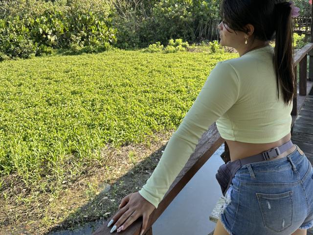 VictoriaWaynes with her back to the camera, wearing a yellow long-sleeve crop top and blue jeans, leaning on a wooden railing by a waterway. Her ass is prominent.