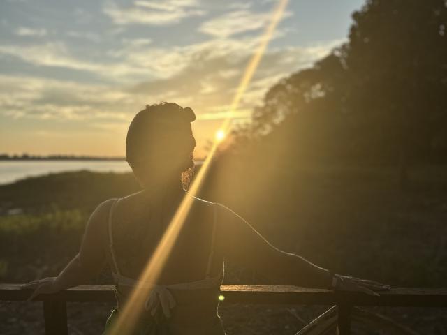 Silhouetted VictoriaWaynes with sun flare, back to camera, arms on wooden railing, wearing bikini, sunset, golden light, tree on right, water in background, peaceful, serene.