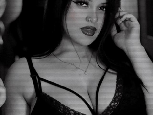 black-and-white photo of CamiDam with dark lipstick, long hair, wearing a lace bra, showing her large tits, hand in hair, necklace, looking seductively at the camera
