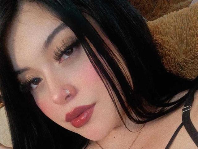 CamiDam has black hair, a nose piercing, and is wearing red lipstick with long eyelashes, showing a slightly pouty expression, with her tits barely visible in the frame.