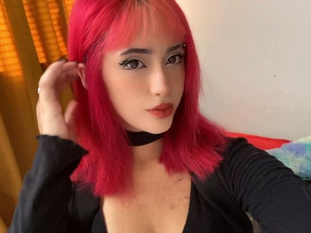 PriscilaLyn with bright pink hair, black choker, and black top, has a nose ring and eyeliner, looking at the camera with a slight smile, holding her hair.