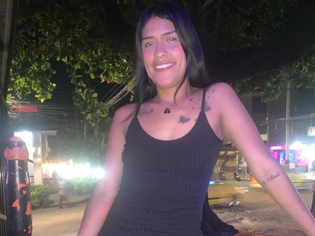 MilaNovak with long black hair, smiling, wearing a black ribbed tank top, showing tattoos on shoulders and chest, standing at night on a street with neon lights.