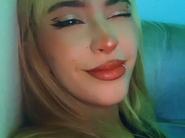 PriscilaLyn's face, winking, with blonde hair, black eyeliner, red glossy lips, nose piercing, and lip ring, smiling slightly, against a greenish-blue background.