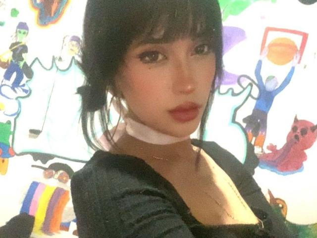 PriscilaLyn with black hair, bangs, and red lipstick, wearing a black top and white choker, showing her tits slightly in a selfie. Background is colorful and abstract.