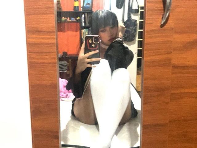 PriscilaLyn sits in front of a mirror, wearing black lingerie and white thigh-high socks, with her knees up, showing her bare ass. She holds a phone, taking a selfie.