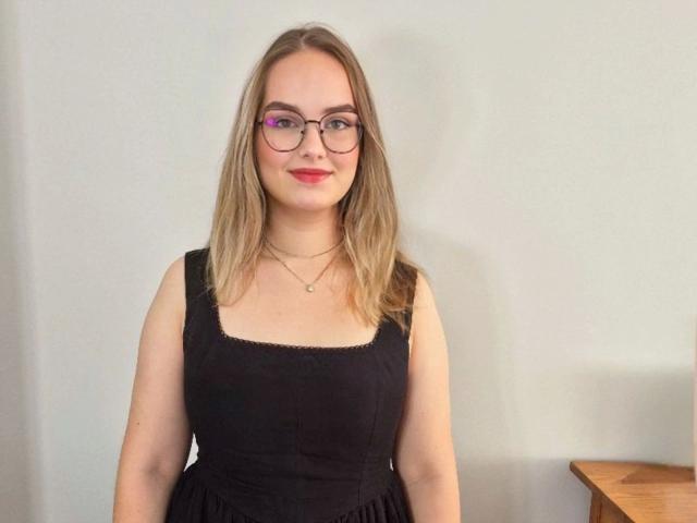 AliceFetish-hot has blonde hair, glasses, and red lipstick, wearing a black dress with a square neckline. She's standing with a slight smile, looking at the camera.