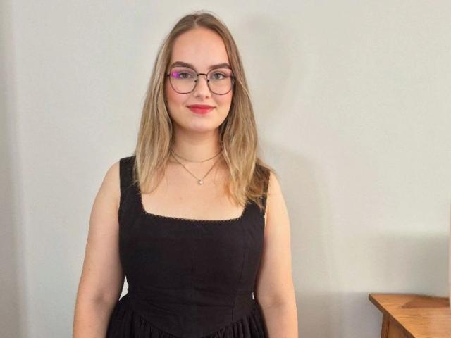 AliceFetish-hot with glasses, red lipstick, and blonde hair, wearing a black dress, standing against a white wall, and a wooden table partially visible on the right.