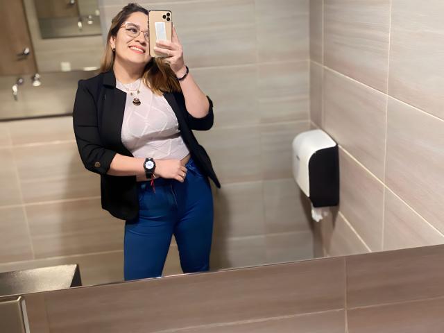 ChloeDlunet met een bril, lachend, gekleed in een zwart blazer, een wit, licht doorschijnend topje, blauwe jeans en een horloge, die een selfie maakt in een badkamer en daarbij haar borsten laat zien..