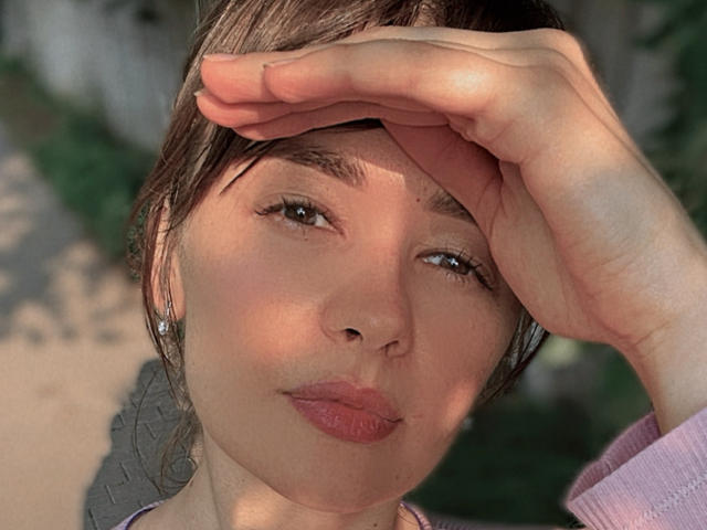 SashaElisa with a hand shielding her eyes, brown hair, light skin, full pink lips, subtle makeup, small earrings, pink shirt, sunlit face, slight smile, soft natural light.