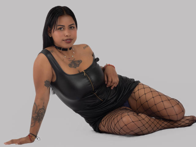 Michy69 with black leather dress, zipper down, fishnet stockings, tattoos on arms and chest, choker, lying on floor, smiling, hand on thigh, dark hair, tan skin.