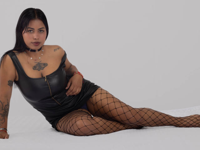 Michy69 is a Latina woman with black hair, wearing a black leather dress, fishnet stockings, and a choker, showing her tits and tattoos, laying on a white surface.