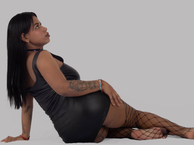 Michy69 with black hair, wearing a tight black leather dress and fishnet stockings, has tattoos on her arm, sitting with her ass up, looking to the side.