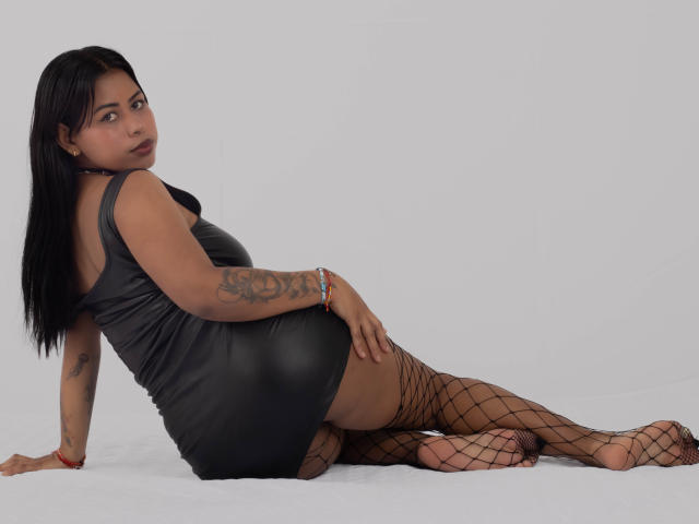 Michy69 with dark hair, wearing a black leather dress, fishnet stockings, and red bracelet, laying on white, looking back, showing tattoo on right arm, and her ass.
