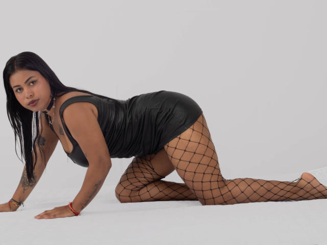 Michy69, dark-haired, wearing a black, shiny, tight, sleeveless dress, and fishnet stockings, is on all fours, looking at the camera, with a tattoo on her right arm.