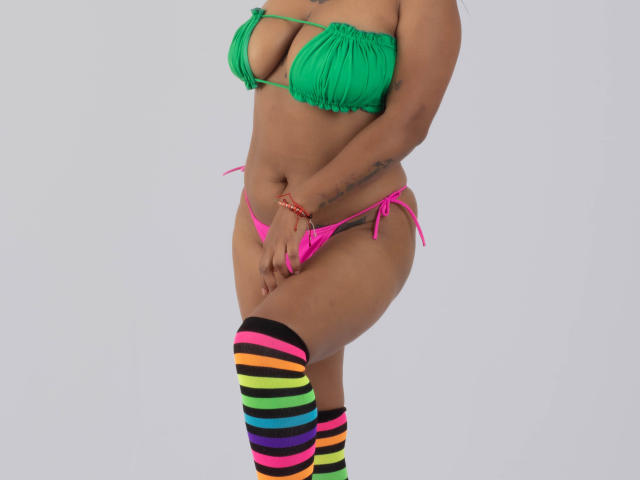 Michy69 with green bikini top showing boobs, pink bikini bottom, and colorful striped thigh-high socks, standing, hands near pussy, tan skin, tattoo on right hip.