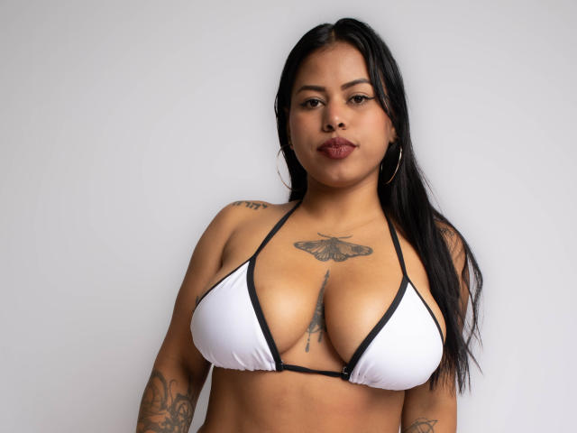 Michy69 with medium brown skin, black hair, and hoop earrings, wearing a white bikini top, showing large tits and a moth tattoo on her chest, and a vertical tattoo below.