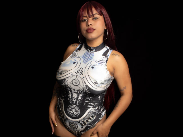 Michy69 with red hair, wearing a high-cut, black-and-white graphic swimsuit, showcasing her big tits and tattoos, standing against a black background, looking confident.