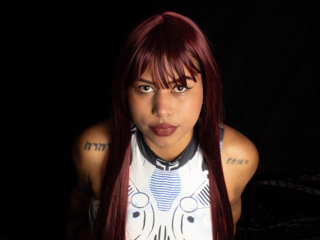 Michy69 with red hair, tattoos on shoulders, wearing a white and blue sleeveless top, looking directly at camera with pouty lips, dark background. Boobs, ass, tits, pussy not visible.