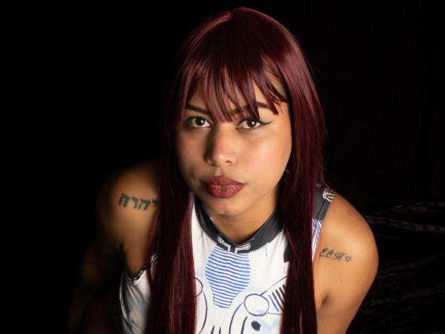 Michy69 with dark red hair, tattoos on shoulders, pouty lips, brown eyes, wearing a white and blue sleeveless top, black background, looking directly at camera.
