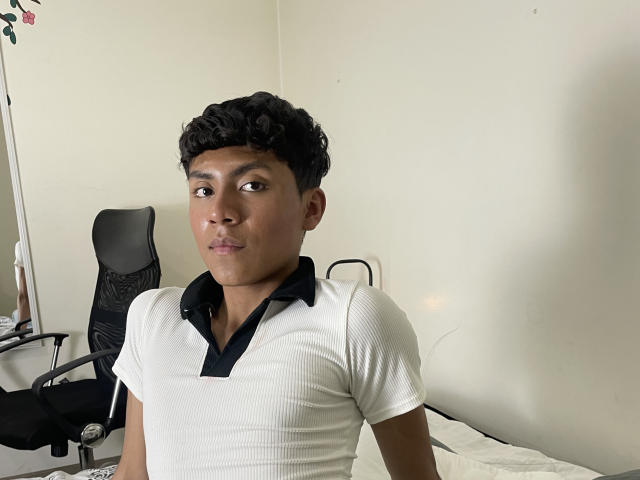 Jhundy69, avec des cheveux noirs bouclés, une peau marron moyen et un t-shirt blanc à rayures avec un col noir, faisant légèrement la moue, dans une pièce simple avec une chaise noire..