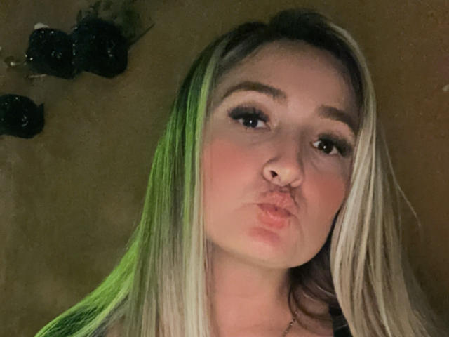 Antonella69X has pouty lips, green and blonde hair, and is giving a puffy kiss face, with dark eyeliner and a slight blush, in a dimly lit photo.
