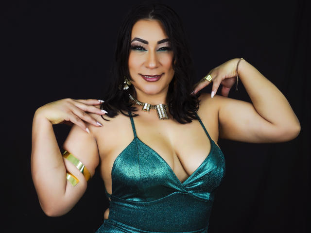 EmaFoxy tiene senos grandes, lleva un vestido verde brillante, un collar dorado y pulseras, tiene el pelo oscuro, está sonriendo y posando con las manos en los hombros sobre un fondo negro..