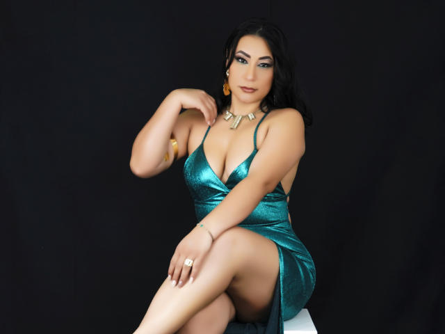 EmaFoxy with big tits, wearing a shiny green dress, sitting with one leg up, showing a thigh, black hair, gold necklace, and ring, against a dark background.