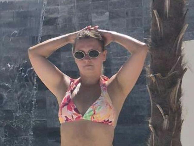 AngryGirl69 stands with arms raised, wearing a colorful floral bikini top, round sunglasses, and short brown hair. She has a medium build and tan skin.